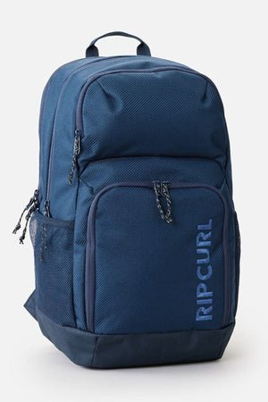 The Rip Curl Chaser 33L Backpack, made from recycled polyester, boasts a trendy blue design with various compartments and zippers, including a front pocket. It securely accommodates a 15" computer and includes side mesh pockets, a top handle, alongside the "RIP CURL" logo printed vertically on the front.