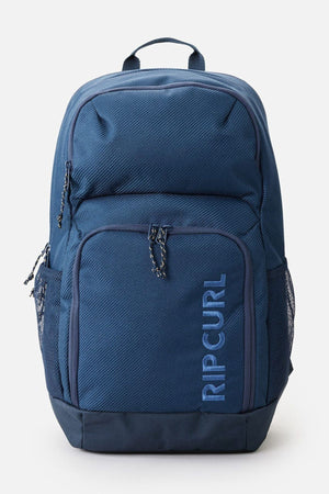 The Rip Curl Chaser 33L Backpack, made from recycled polyester, showcases a blue color with a front pocket, dual side mesh pockets, and safe zippered compartments. The brand name is prominently displayed vertically on the front. It features a textured finish and includes a top carrying handle.