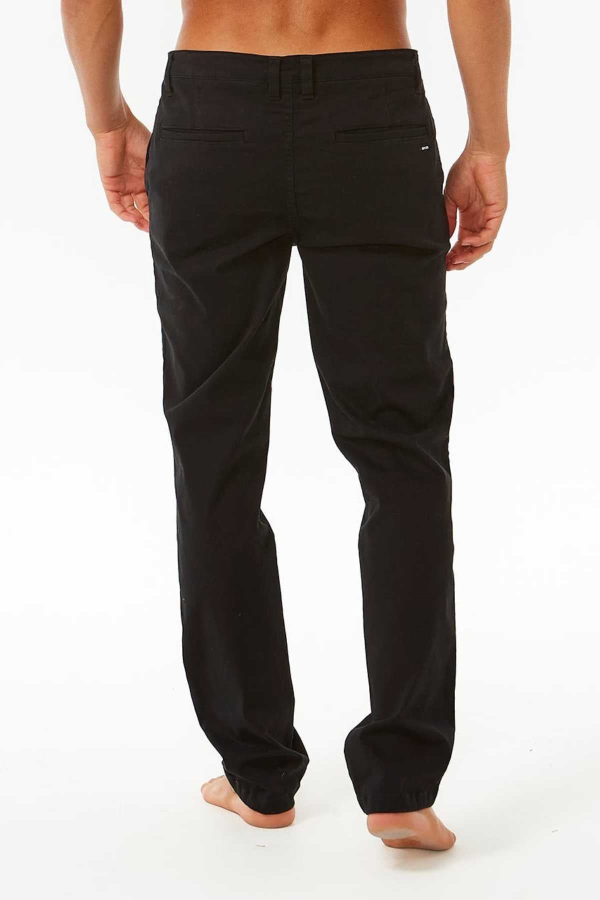 The person stands barefoot, facing away from the camera in sleek black Rip Curl Classic Surf Chino pants made of stretch cotton twill, set against a plain white backdrop that highlights their effortless style.