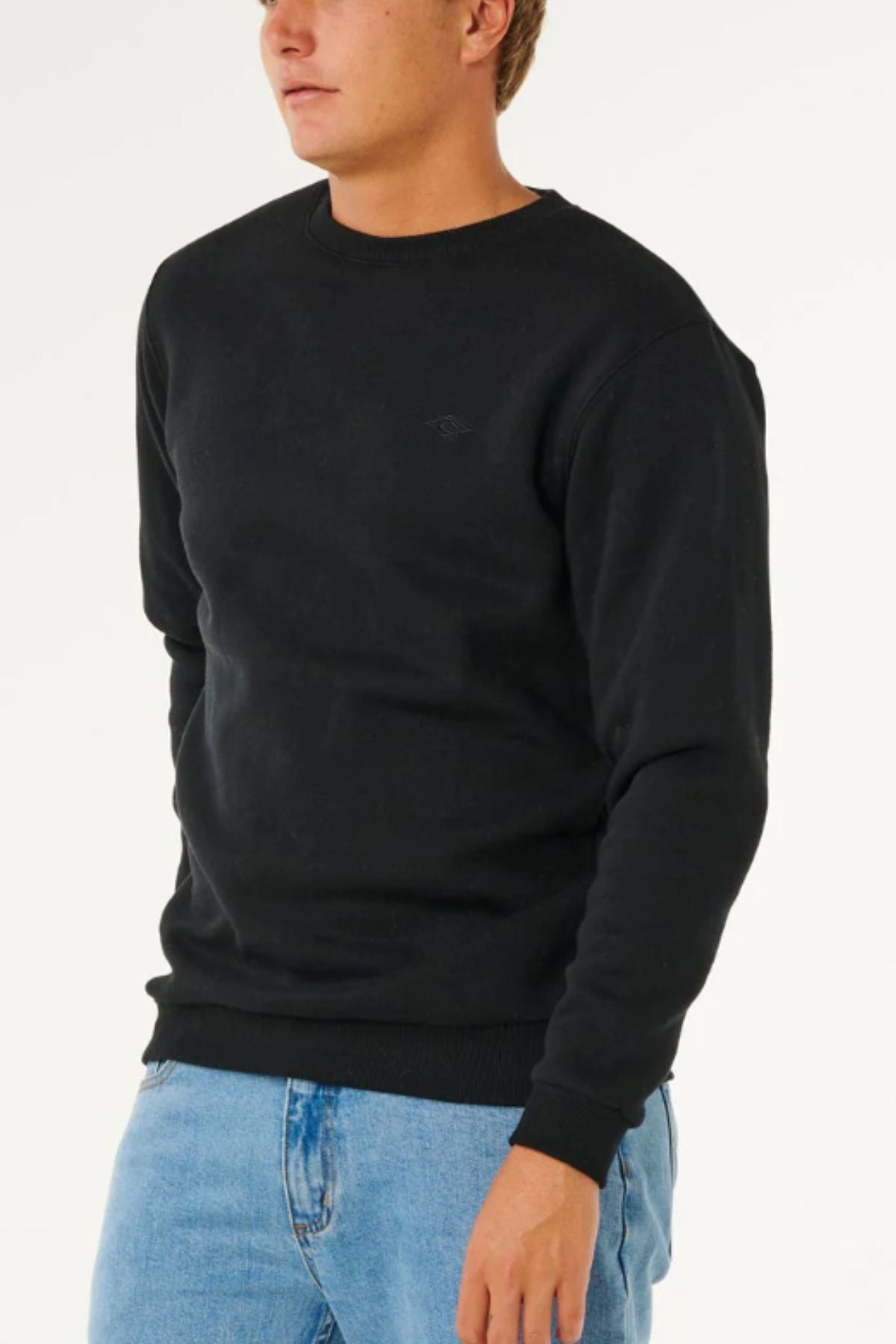 Wearing the Rip Curl Classic Surf Crew black sweatshirt with an embroidered logo and light blue jeans, a person stands against a white background, partially out of frame to highlight this surf essential style.
