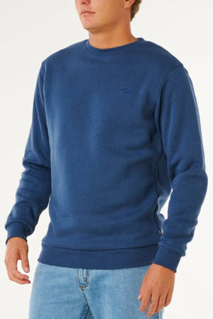 A man wears a Rip Curl Classic Surf Crew blue sweatshirt with an embroidered logo and light blue jeans, standing against a white background for a classic surf-inspired look.