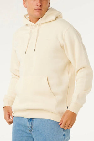 A person wearing the Rip Curl Classic Surf Hood, a cream-colored hoodie with a front pocket, and blue jeans stands against a plain light background.