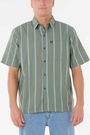 A person wears a Rip Curl Classic Surf Stripe Short Sleeve Shirt—green with light blue vertical stripes and a chest pocket—paired with light blue jeans, standing against a plain white background.