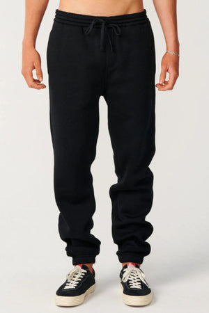 Someone stands against a plain light background, chest down visible, wearing Rip Curl Classic Surf Trackpant in black with a relaxed fit and drawstring waist, paired with black-and-white sneakers—ideal for casual wear.