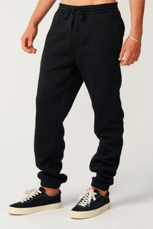 Someone wearing the Rip Curl Classic Surf Trackpant with a drawstring waist and black sneakers with white soles stands against a plain light background—ideal for relaxed, easygoing casual wear.