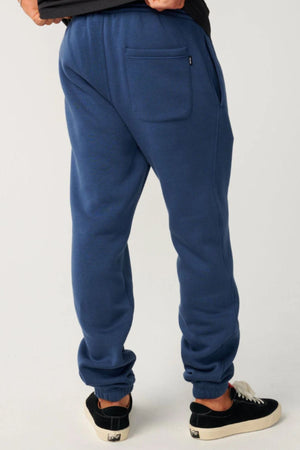 A person in relaxed fit Rip Curl Classic Surf Trackpant and black sneakers faces away, highlighting the back pocket and elastic cuffs against a plain light background.