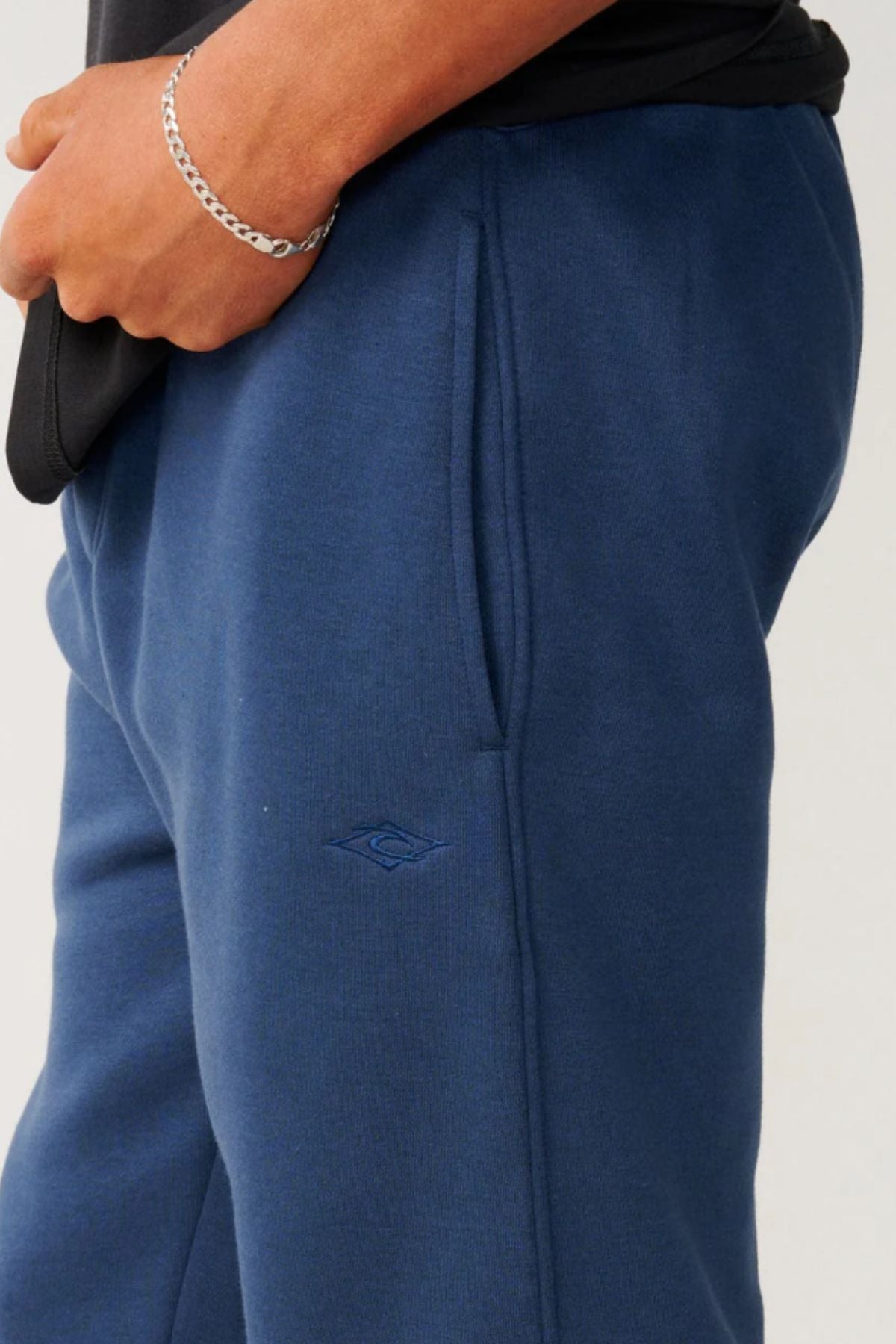 A person wears Rip Curl Classic Surf Trackpant in dark blue with a relaxed fit, right hand in the pocket showing a silver chain bracelet. The sweatpants feature a small embroidered logo near the pocket, ideal for casual wear.