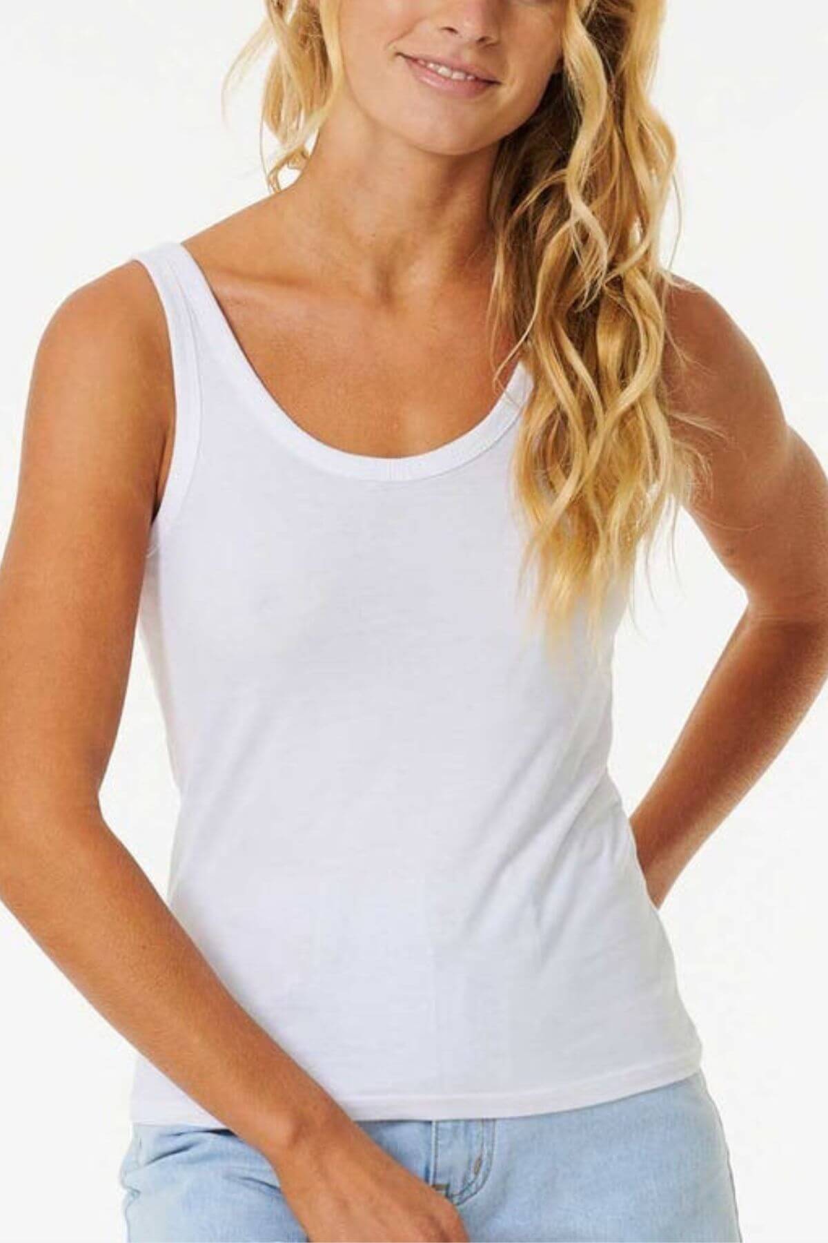 Woman with long blonde wavy hair wears the Rip Curl Classic Tank in white cotton jersey and light blue jeans, standing against a plain background and smiling gently—the ideal layering piece for any casual style.