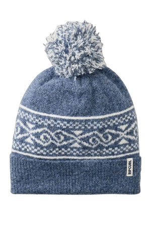 The Rip Curl Cosmic Kiss Jacquard Beanie is blue with a white geometric pattern, folded brim, fabric "RIP CURL" tag, and a large pom-pom—ideal for stylish winter wear.