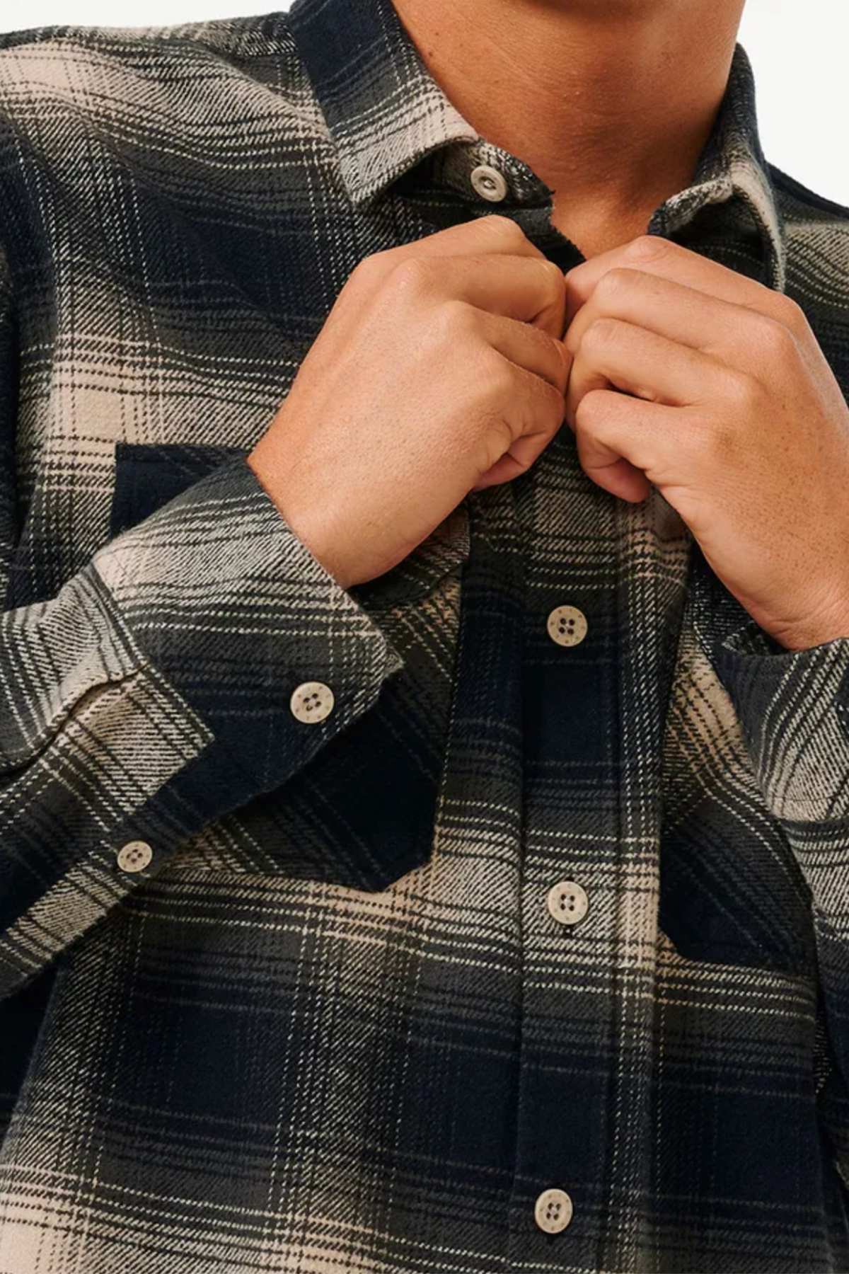 A person buttons up the Rip Curl Count Flannel Shirt, featuring soft cotton flannel with a garment-wash finish. Only their torso and hands are shown, emphasizing the dark plaid design with beige and navy stripes.