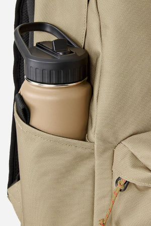 A tan water bottle with a black lid sits in the side pocket of a beige Rip Curl Crusha 30L Search Backpack made from durable fabric.