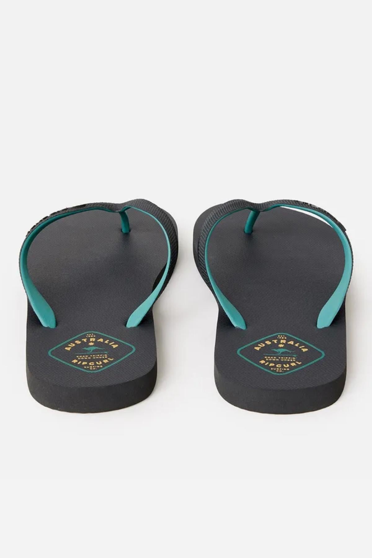 A pair of Rip Curl Desto Bloom Thongs featuring black flip-flops with teal straps. The footbed has a small teal and gold emblem on the heel area, set against a plain white surface.