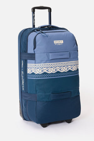 The Rip Curl F-Light Global 100L Cosmic Kiss Wheeled Travel Bag features blue and navy tones, white geometric and wave patterns, a retractable handle, wheels, and AirTag compatibility for added convenience.