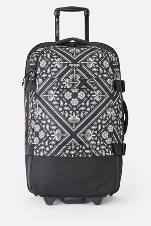 The Rip Curl F-Light Global 100L Bossa Nova is a black check-in roller bag with a white paisley and geometric pattern, telescopic handle, side straps, black bottom, AirTag sleeve, and the Rip Curl logo near the top front.