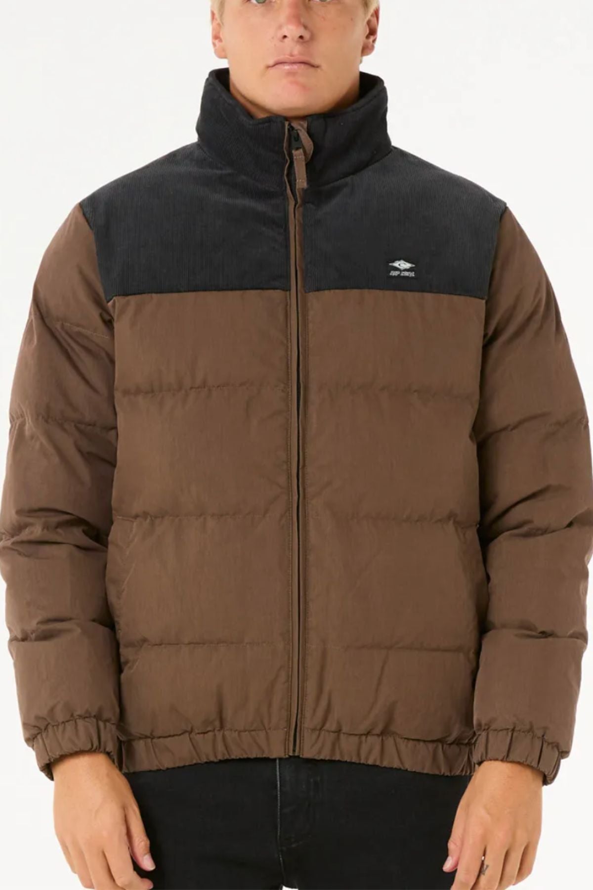 Someone stands against a plain white background wearing the Rip Curl Fade Out Cord Puffer Jacket: a brown cord jacket with a dark upper panel, high collar, quilted padding, and small left-chest logo—ideal for outdoor adventure.