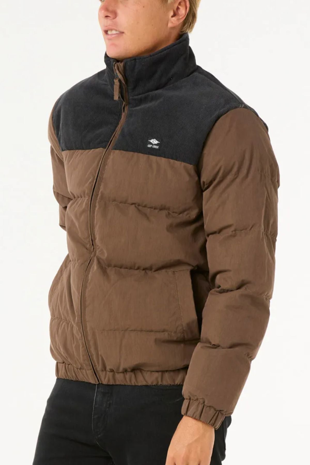 Someone stands against a plain light background wearing the Rip Curl Fade Out Cord Puffer Jacket—brown with dark gray shoulders and collar, quilted padding, and a zip-up front—paired with black pants.