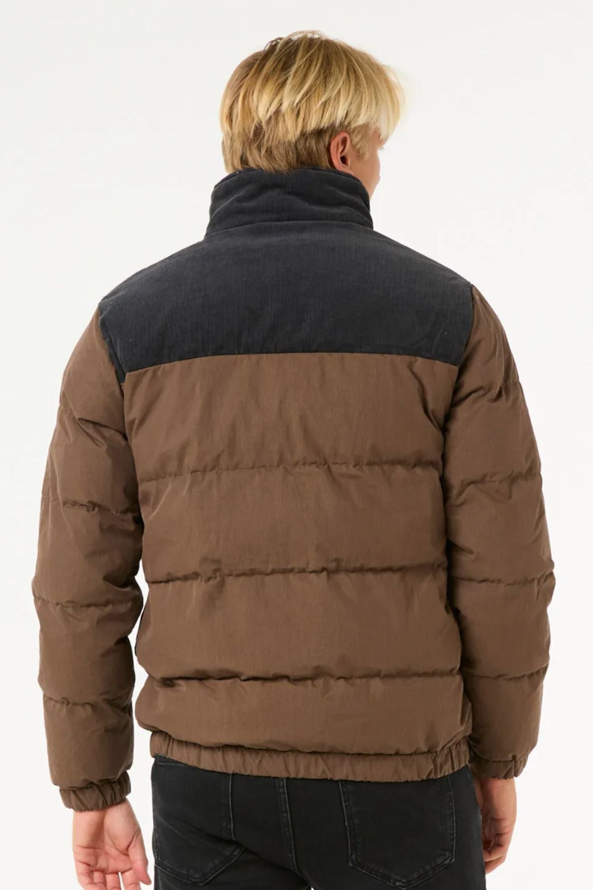 A person with blonde hair, seen from behind, is wearing the Rip Curl Fade Out Cord Puffer Jacket in brown with quilted padding and a dark gray yoke, paired with black pants against a plain white background.