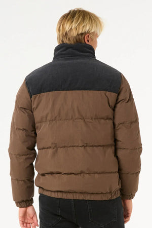 A person with blonde hair, seen from behind, is wearing the Rip Curl Fade Out Cord Puffer Jacket in brown with quilted padding and a dark gray yoke, paired with black pants against a plain white background.