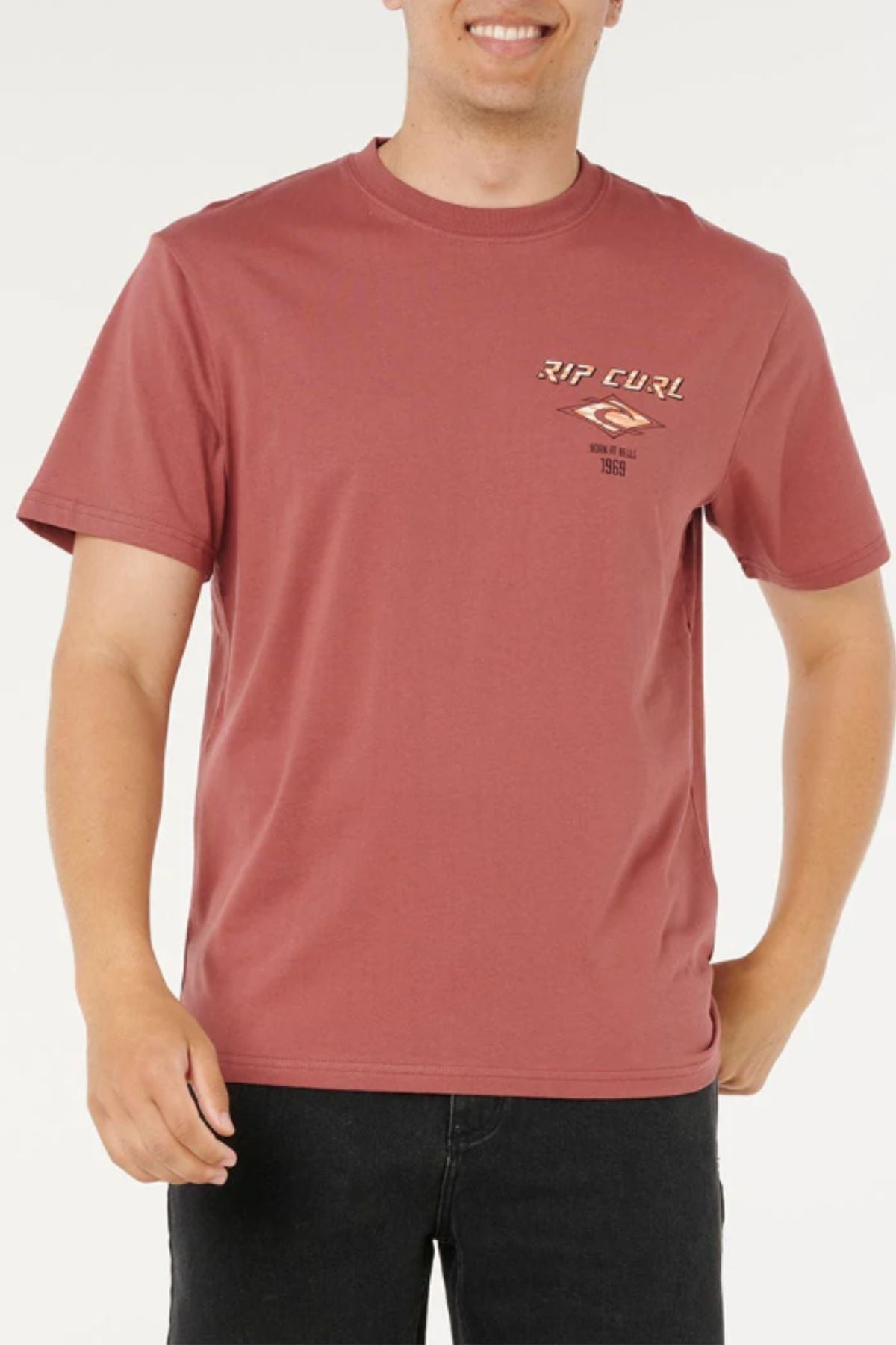 Someone in a faded red Rip Curl Fade Out Icon Tee, a men's cotton t-shirt with a small left chest logo, paired with black jeans, stands against a plain white background.