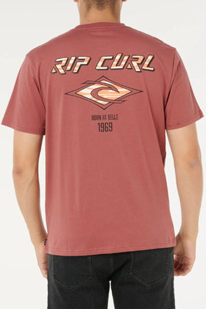 A person faces away from the camera wearing the Rip Curl Fade Out Icon Tee, a mauve men’s cotton t-shirt featuring “RIP CURL,” a wave logo, and “Born at Bells 1969” on the back.