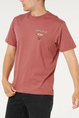 Wearing the Rip Curl Fade Out Icon Tee, a classic faded red men’s shirt featuring a chest logo, paired with black jeans, the person stands against a white background looking to the side.