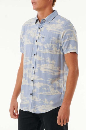 An individual is wearing the Rip Curl Fun Times Short Sleeve Shirt, a breathable light blue shirt adorned with a white tropical palm tree pattern. This short-sleeved button-up shirt includes a pocket on the left chest featuring a small black tag, and the person is facing slightly to the right.