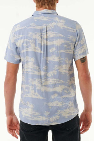 A person is dressed in a Rip Curl Fun Times Short Sleeve Shirt, which is light blue and features a white tropical palm tree design. This breathable shirt complements the person's dark pants perfectly. The image shows them from behind against a plain white backdrop.