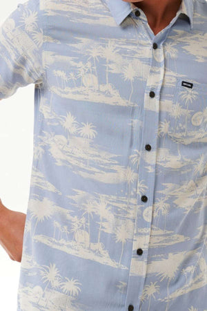 A person wears the Rip Curl Fun Times Short Sleeve Shirt, which is light blue and adorned with a white tropical island pattern featuring palm trees. This breathable shirt also includes a small chest pocket with a label. Only part of the person's torso and arm is visible.