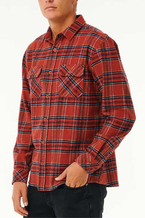 A person dressed in a Rip Curl Griffin Flannel Shirt, characterized by its long sleeves and red plaid pattern with blue and white stripes. The shirt, crafted from heavy brushed fabric, features two chest pockets. They have one hand in their pocket and wear black pants, standing against a plain background.