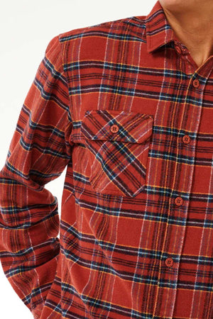 Close-up of an individual wearing the Rip Curl Griffin Flannel Shirt, showcasing its red and black plaid design. The heavy brushed fabric ensures both comfort and warmth. The shirt includes a single chest pocket on the left side with a diagonal plaid pattern and is buttoned up, displaying only the upper torso alongside part of the neck and chin.