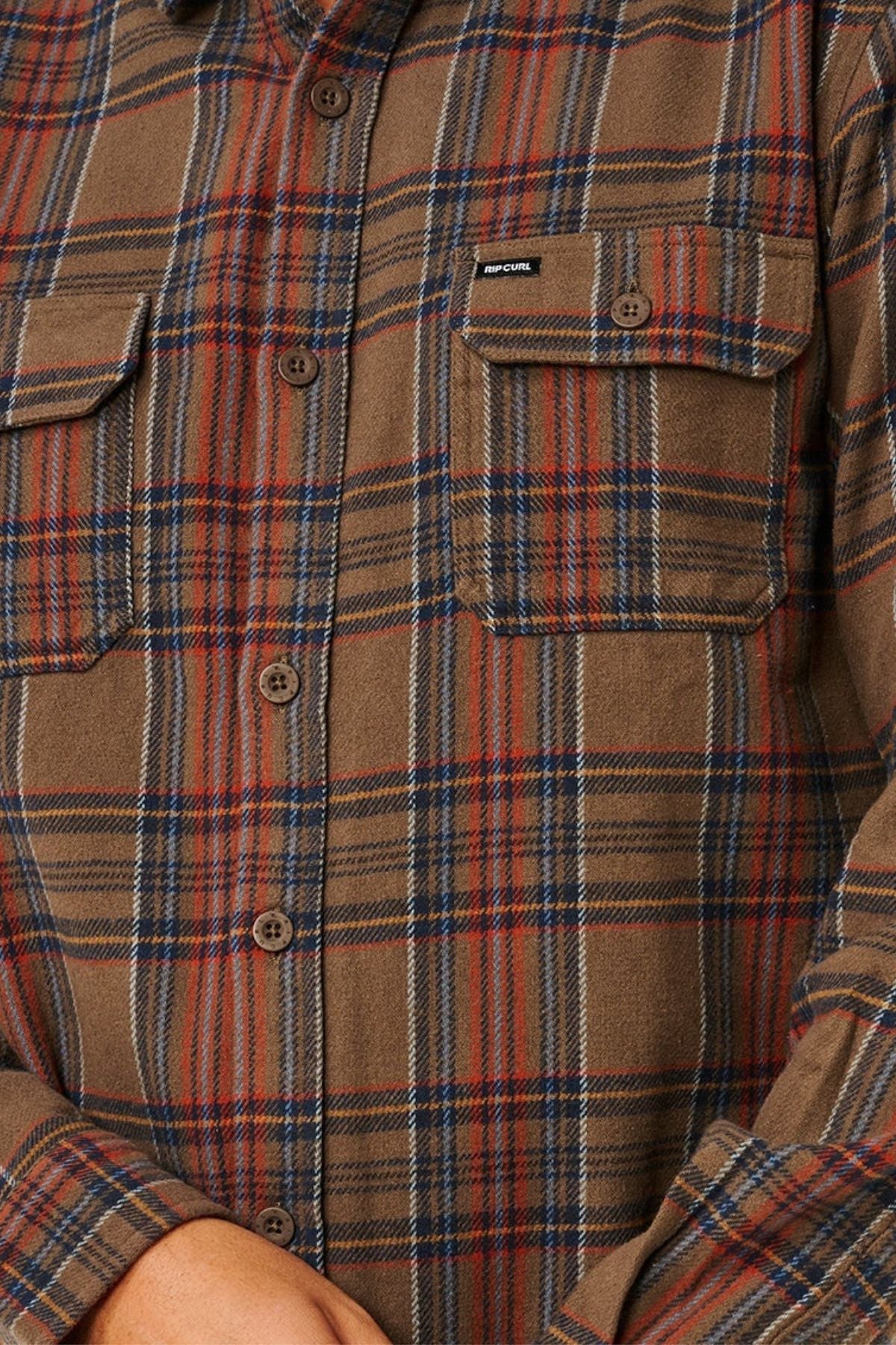 A close-up of a person wearing the Rip Curl Griffin Flannel Shirt in brown, red, and blue plaid with buttoned chest pockets. The person's hands are partially visible at the bottom of the image.