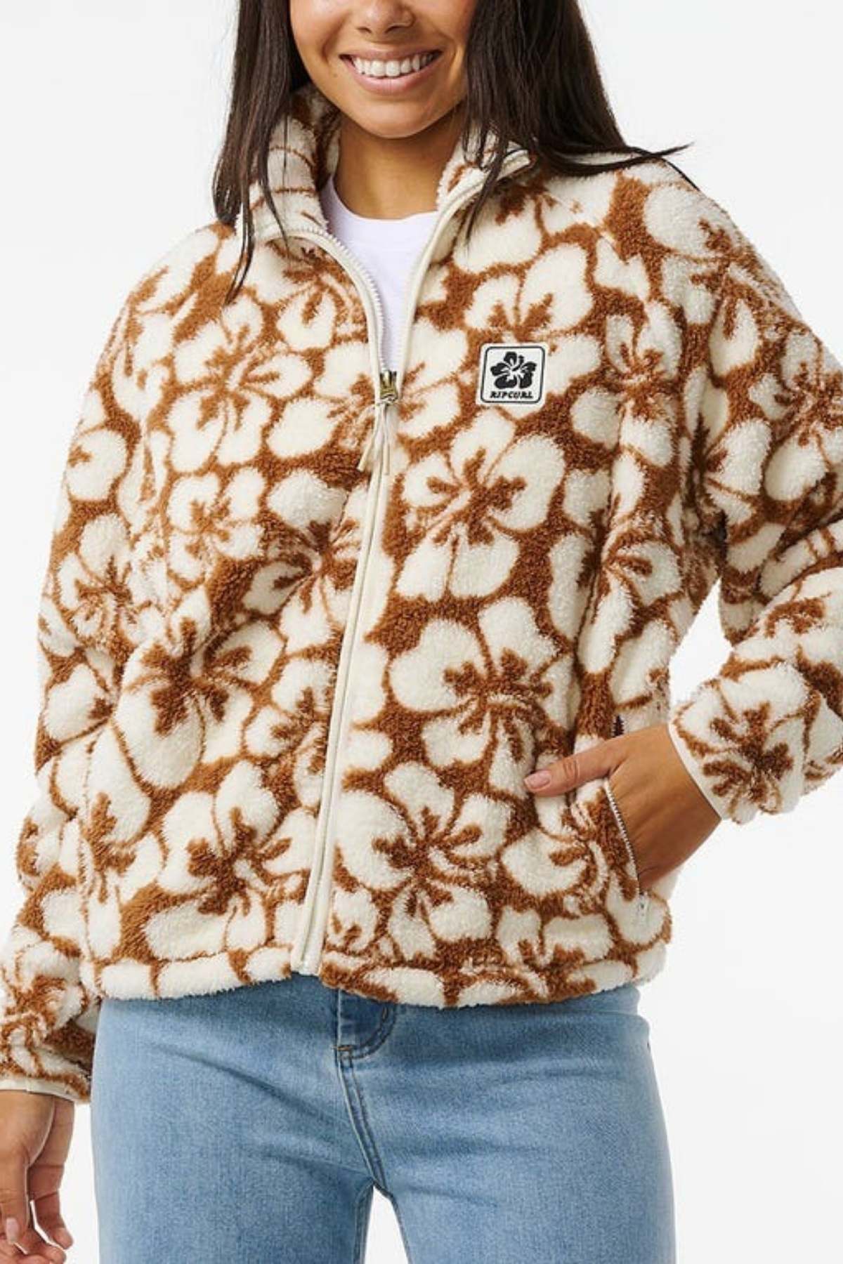 A person is wearing the Rip Curl Hibiscus Heat Polar Fleece Zip Through jacket in brown and white floral, paired with light blue jeans. The jacket has a black and white chest patch, and they smile with their hands in the pockets.