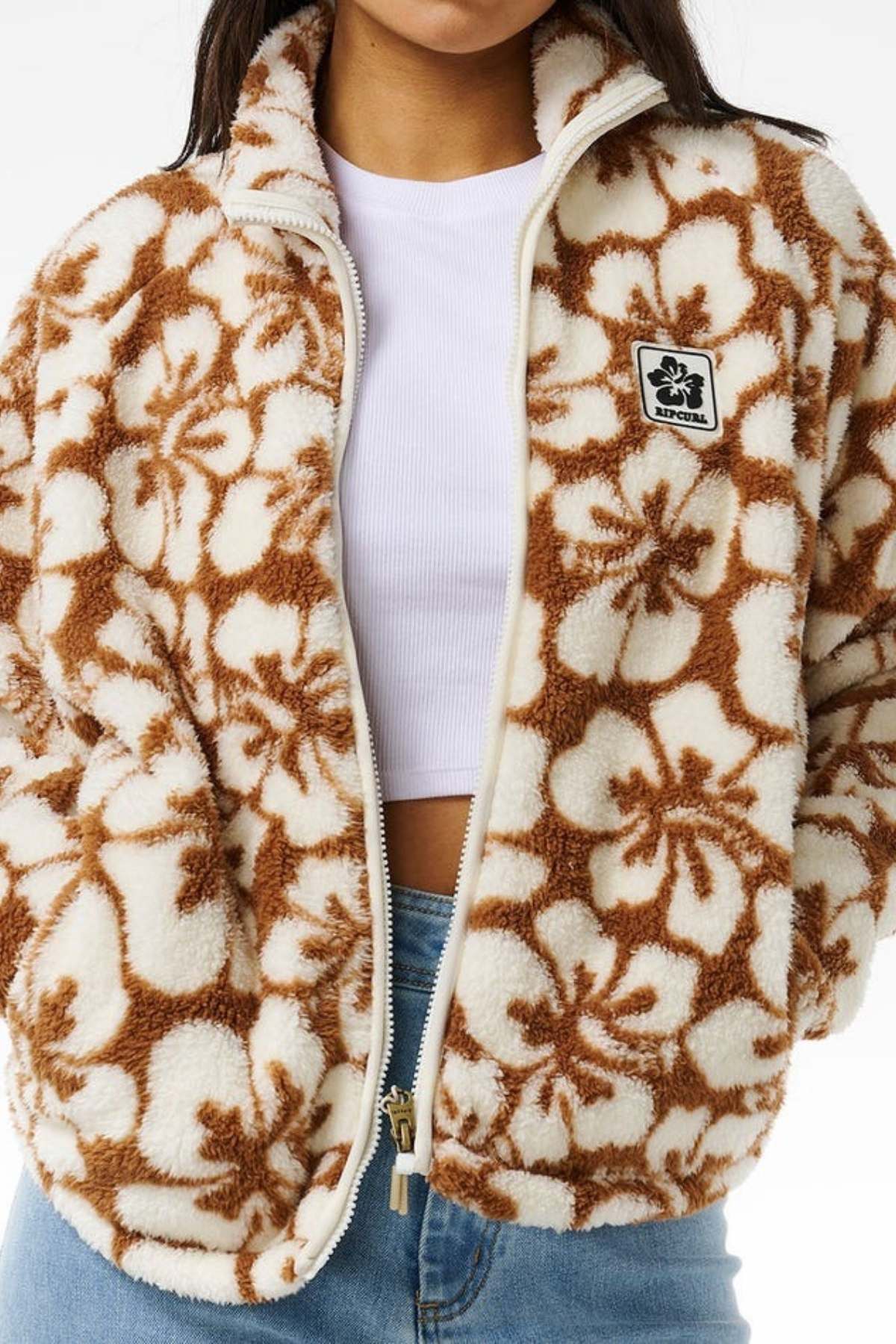 A person wears the Rip Curl Hibiscus Heat Polar Fleece Zip Through, featuring a cream and brown floral pattern and a small Rip Curl patch on the left. They pair it with a white top and light blue jeans against a plain white background.