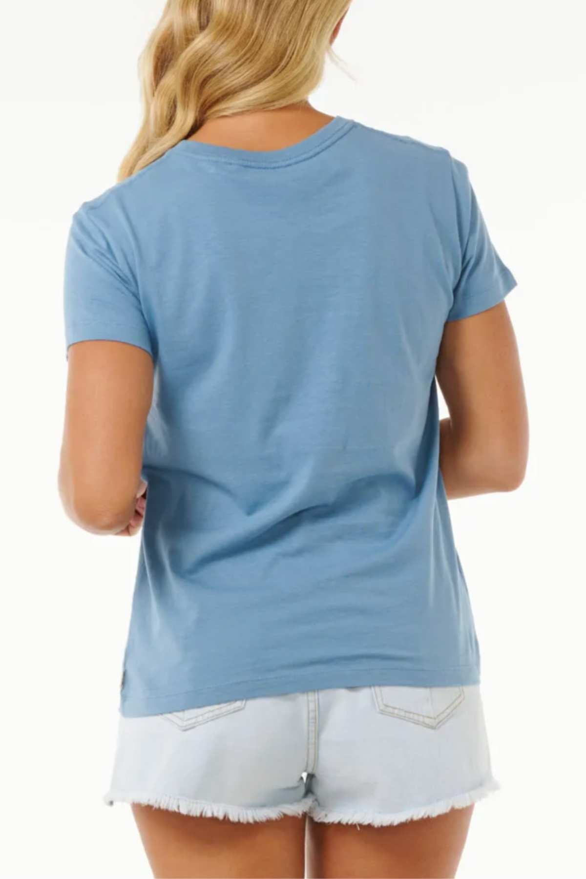 A person with long blonde hair, shown from the back, wears a light blue Rip Curl High Tide Psychic Standard Tee made of soft cotton jersey fabric paired with denim shorts featuring frayed hems. The background is plain and white, highlighting the clothing.