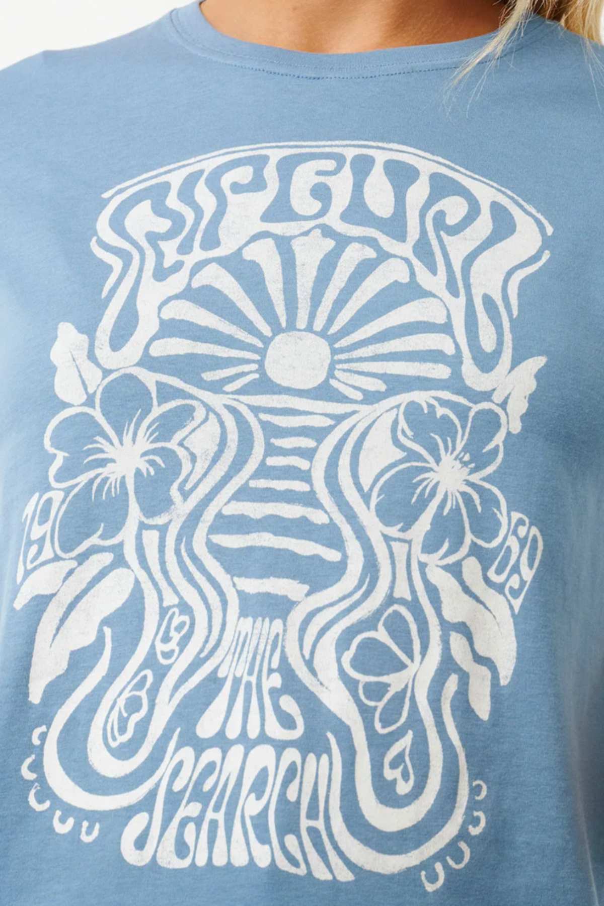 The Rip Curl High Tide Psychic Standard Tee is a light blue cotton jersey fabric shirt featuring a stylized white graphic design with the text "Rip Curl 1969 The Search." The design includes a sun with rays, waves, two hibiscus flowers, and various flowing lines, creating an artistic and surf-inspired look similar to those found in the Rip Curl High Tide collection.