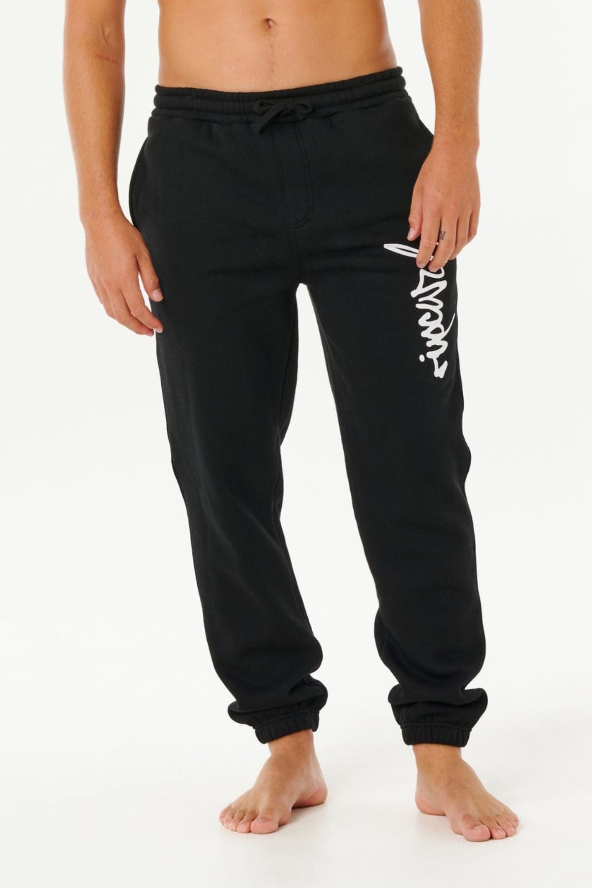 A person wearing Rip Curl Icons Of Surf Trackpant with a white graphic design on the left thigh, made from soft organic cotton fleece, stands barefoot against a plain white background.