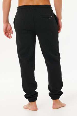 A person stands barefoot on a white background, shown from the back, wearing black Rip Curl Icons Of Surf Trackpant joggers made from soft organic cotton fleece with an elastic waistband, cuffs, and a right-side back pocket.