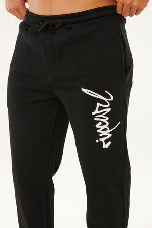 A person wears black Rip Curl Icons Of Surf Trackpant featuring a white stylized script logo on the left thigh, standing against a plain white background.