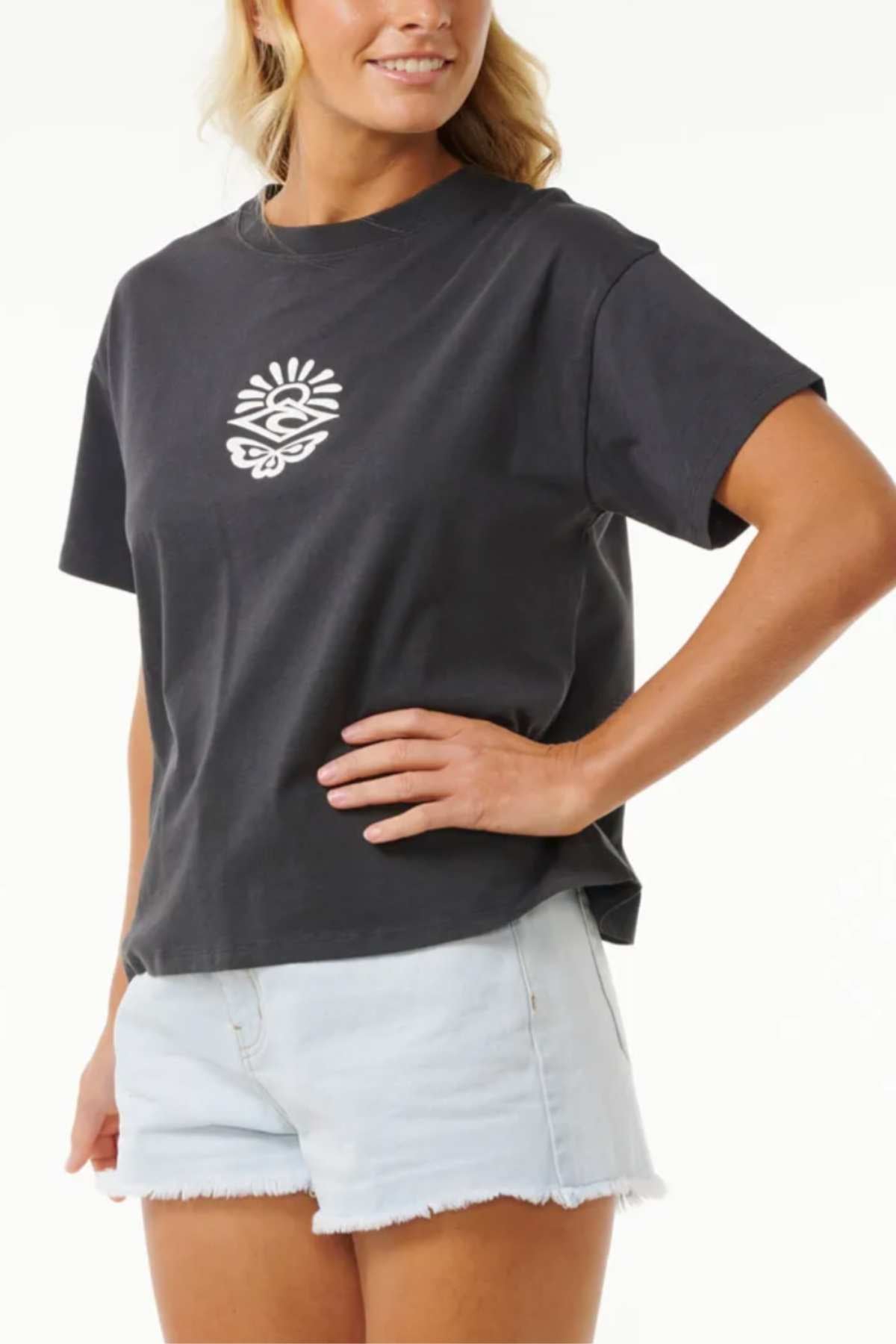A person with long hair is wearing the Rip Curl Icons Of Surf Relaxed Tee, crafted from comfortable organic cotton jersey, paired with light denim shorts. Their hand rests on their hip as they smile slightly against a plain white background.