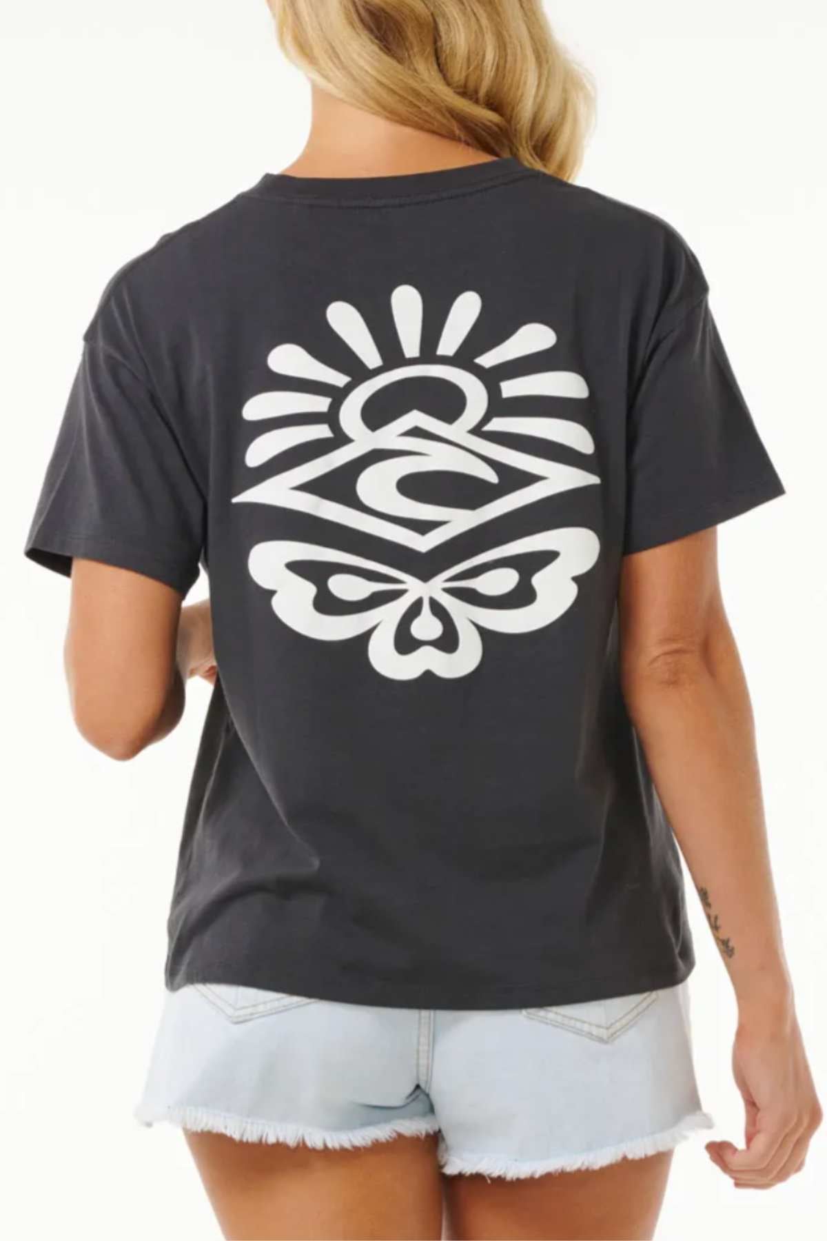 A person with long blonde hair is donning a black Rip Curl Icons Of Surf Relaxed Tee, crafted from organic cotton jersey. The tee boasts a white graphic design featuring abstract shapes and symbols on the back. They have paired it with light-colored, distressed denim shorts against a plain white background.