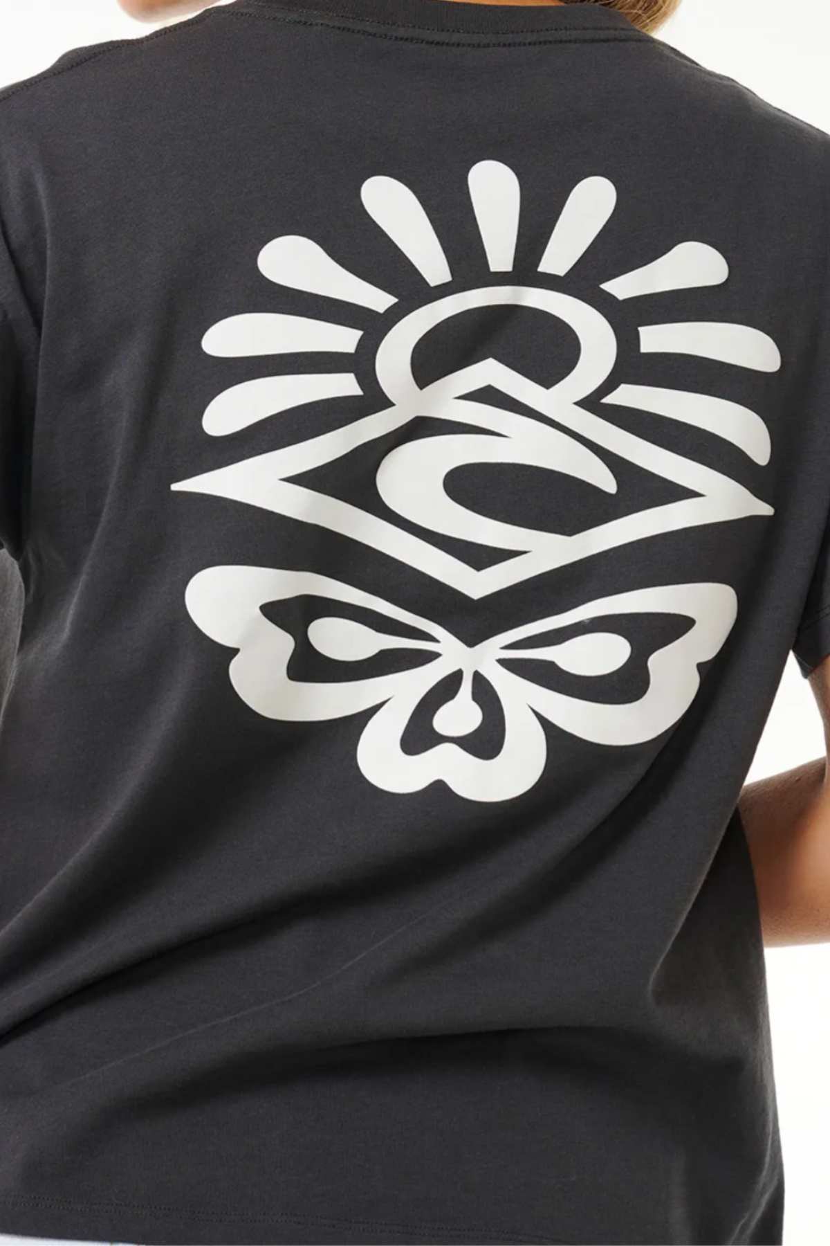 A person wearing a black Rip Curl Icons Of Surf Relaxed Tee made from organic cotton jersey, showcasing a large white graphic on the back with a geometric design that includes a stylized eye, sun rays, and a butterfly at the bottom. The image is captured from behind, highlighting the sustainable style.