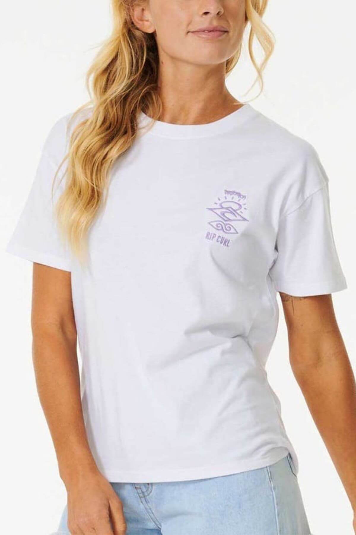 A woman models the Rip Curl Icons Of Surf Relaxed Tee—an organic cotton white short-sleeve shirt with a small purple "RIP CURL" graphic—paired with light blue jeans, standing against a white background in surf-inspired style.