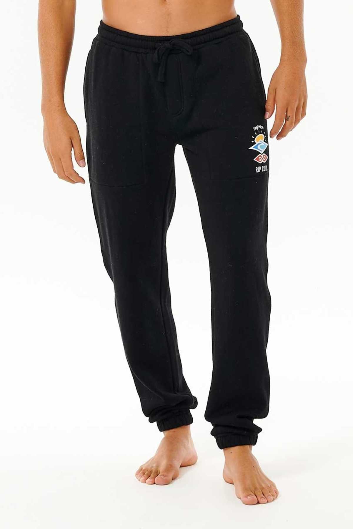 Against a white backdrop, an individual stands barefoot wearing the Rip Curl Icons Of Surf Trackpant, crafted from organic cotton polyester fleece. These black sweatpants feature a drawstring waist and display a small graphic print on the left thigh that includes a cartoon character and the text "RIP CITY." The person's torso is not shown.
