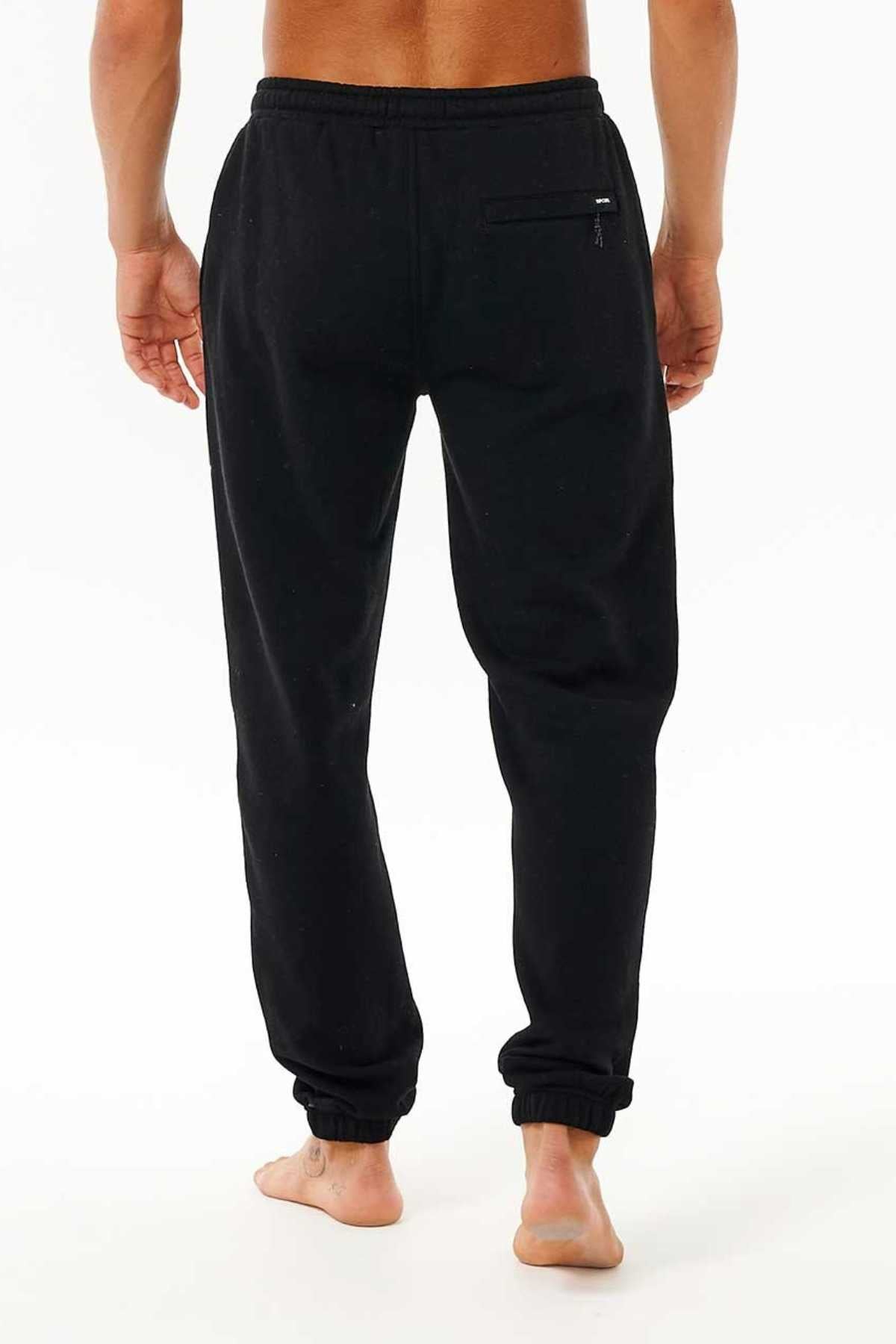 Against a plain white background, a person stands barefoot facing away, wearing the Rip Curl Icons Of Surf Trackpant in black. These joggers are made from Organic Cotton Polyester Fleece and feature a small zippered pocket on the back right side as well as elastic cuffs at the ankles.