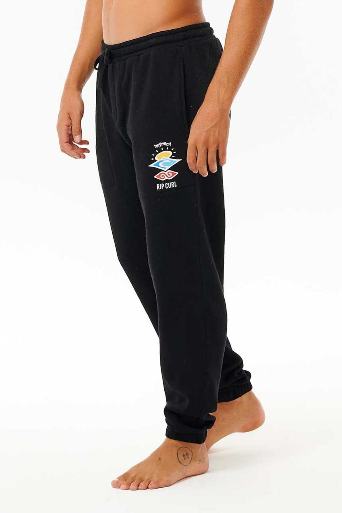 A person wearing the Rip Curl Icons Of Surf Trackpant in black, featuring a drawstring waist, standing barefoot. Made from sustainable comfort organic cotton polyester fleece, the sweatpants showcase a colorful logo on the left thigh with an artistic design and "RIP CURL" text underneath. The individual is positioned against a white background.
