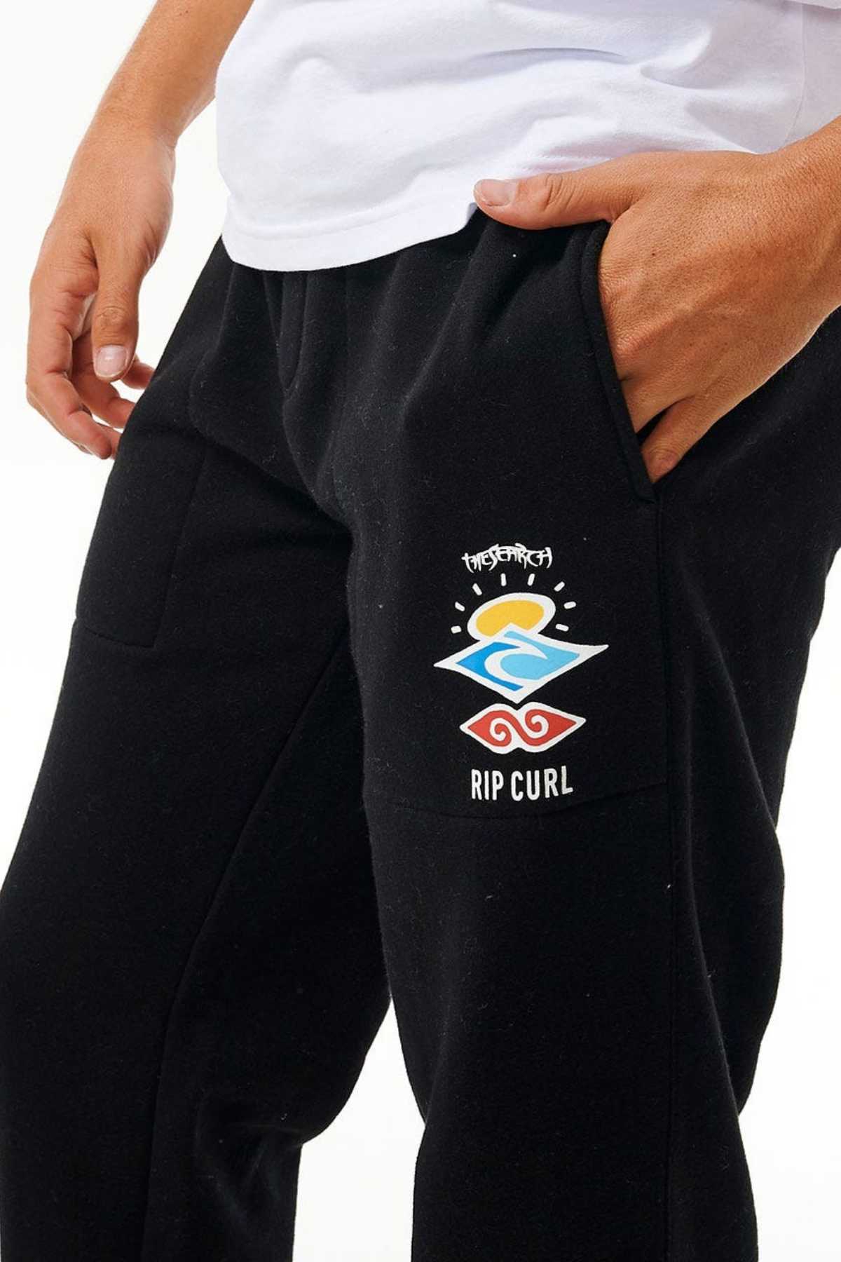 A person wearing Rip Curl Icons Of Surf Trackpants in black, adorned with a vibrant logo on the pocket that showcases a sun, wave, and the word "SEARCH" above the wave. Crafted from sustainable organic cotton polyester fleece for comfort, they also sport a white shirt with one hand tucked casually in the pocket.