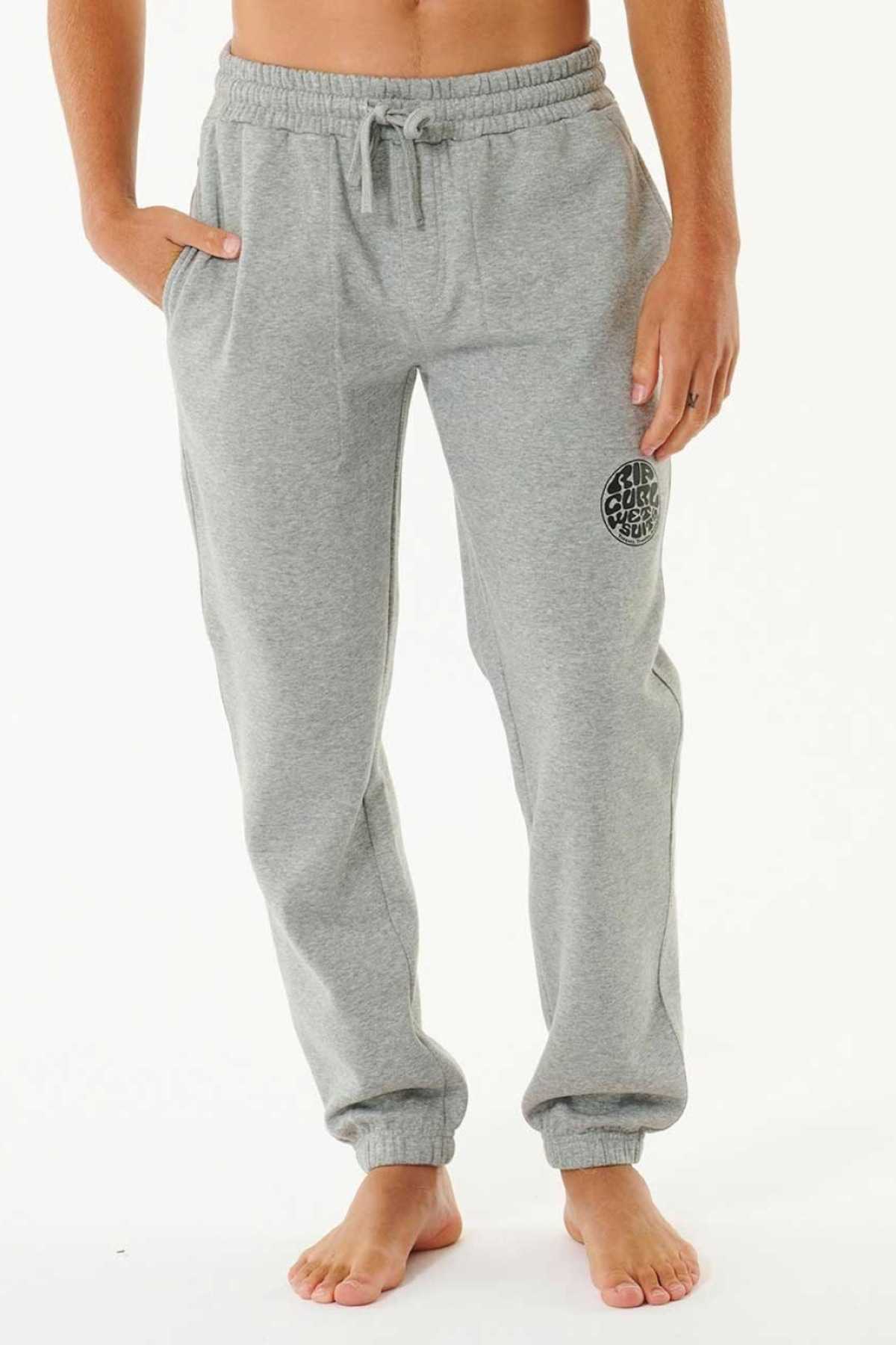 Barefoot against a plain background, a person wears Rip Curl Icons Of Surf Trackpant in light gray organic cotton with an elastic waistband, drawstring, side pockets, and a circular black logo on the upper left leg.