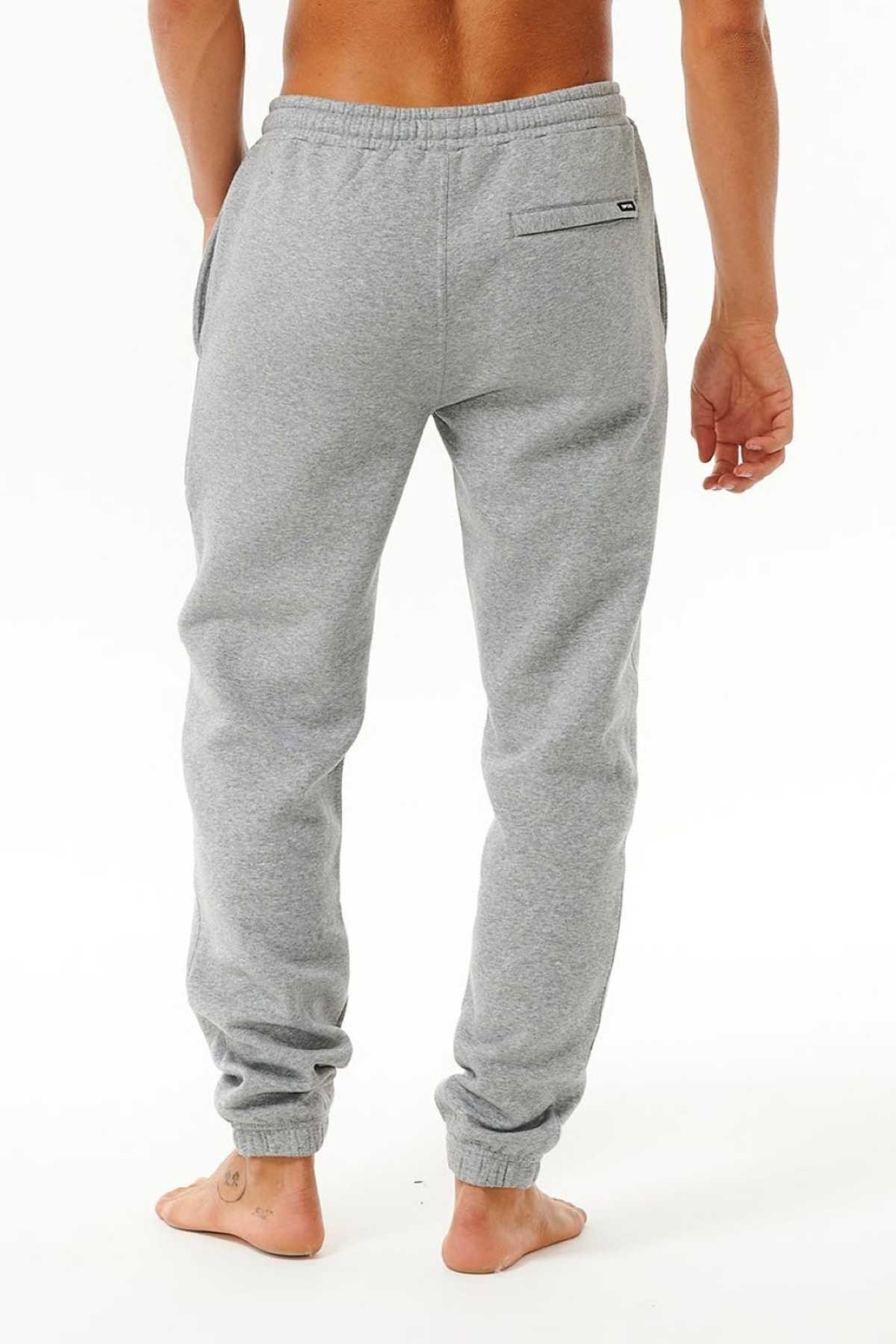 A barefoot person stands on a white background, viewed from the back, wearing light gray Rip Curl Icons Of Surf Trackpant with an elastic waistband and cuffs.