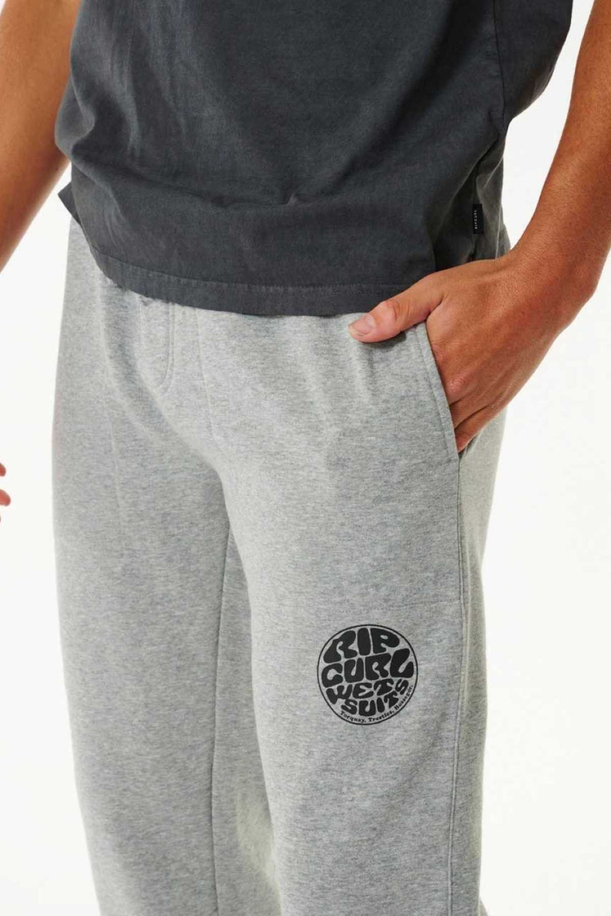 A person wearing a dark gray t-shirt and Rip Curl Icons Of Surf Trackpant made from organic cotton, with one hand in the pocket; the trackpant features a bold round logo graphic on the upper left thigh.