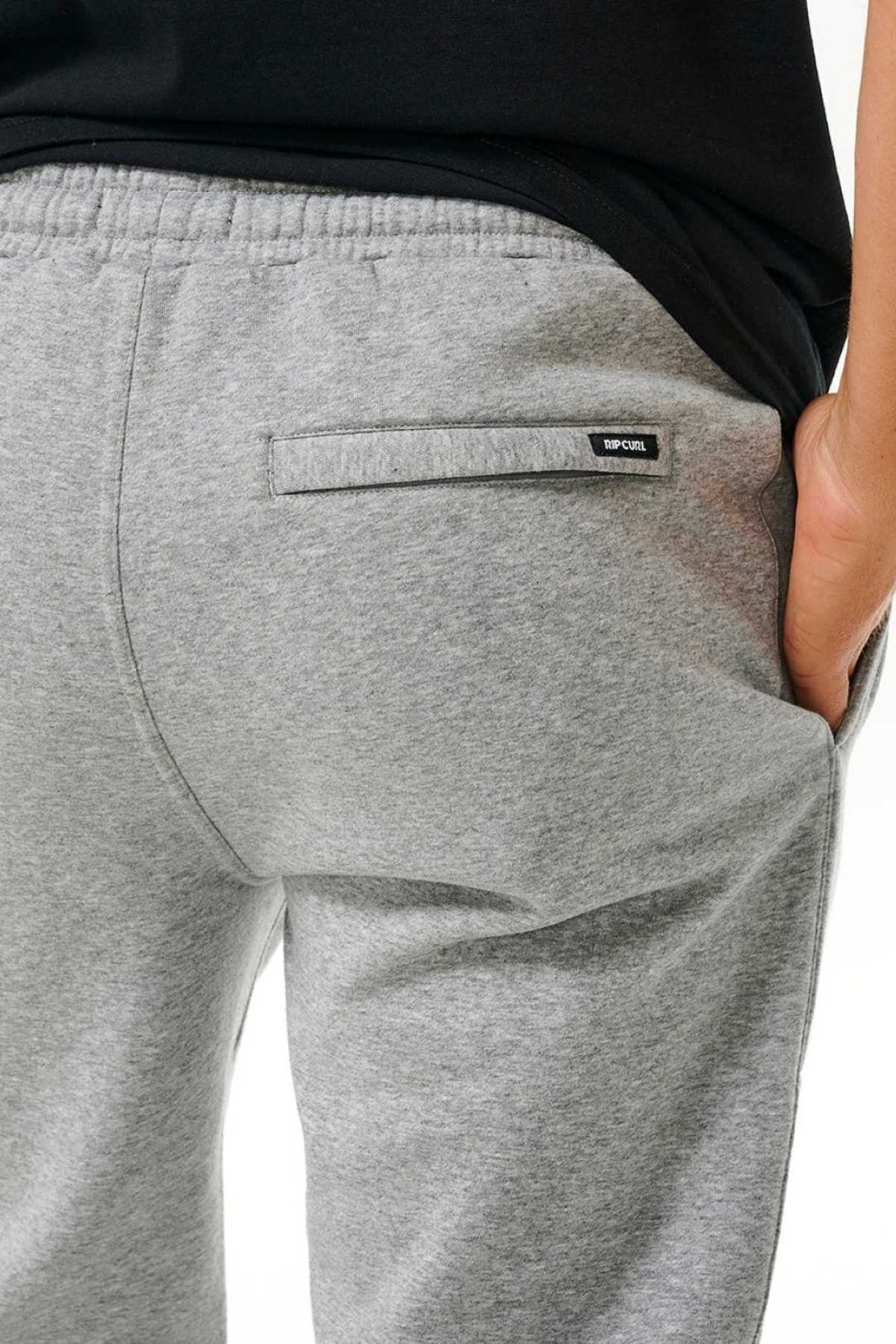 Close-up of a person wearing light gray Rip Curl Icons Of Surf Trackpant, featuring an elastic waistband and black label above the back pocket. Made from organic cotton, these sweatpants are styled with a black shirt and hand in the pocket.